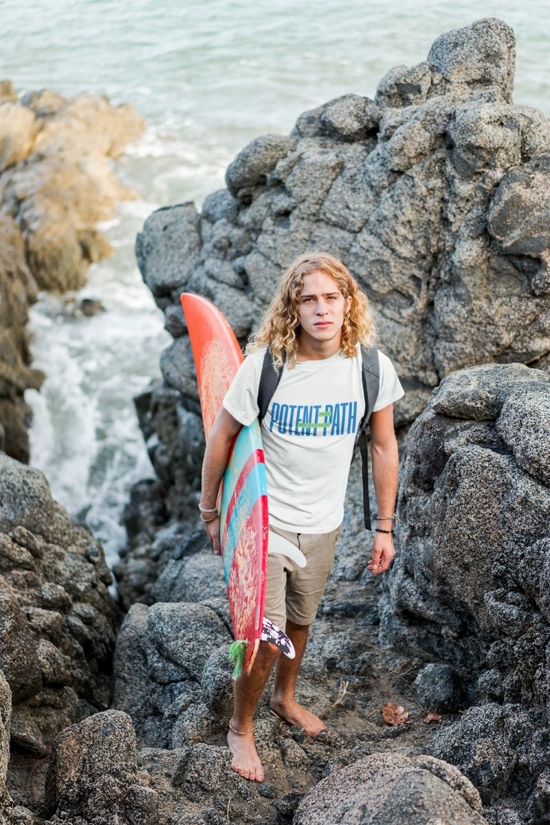 A barefoot man with long blonde hair standing on rocky terrain near the ocean, holding a surfboard and wearing a white Potent Path T-shirt and plain beige shorts, with waves and rocks in the background