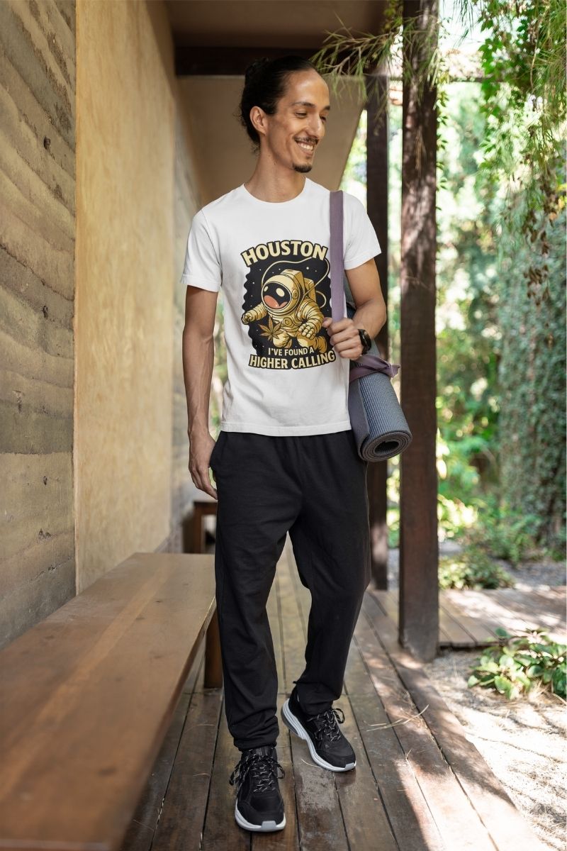 A smiling man with long hair tied back walking along a wooden path wearing a white graphic T-shirt, black pants, and sneakers, carrying a rolled yoga mat over his shoulder in an outdoor, natural setting