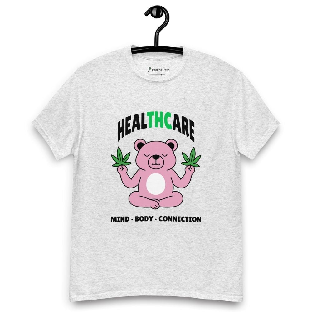 Ash grey T-shirt on a hanger featuring a pink cartoon bear sitting in a meditative pose and holding two cannabis leaves, with the word "HEALTHCARE" above, with the words "THC" highlighted in green and the phrase "MIND, BODY, CONNECTION" below