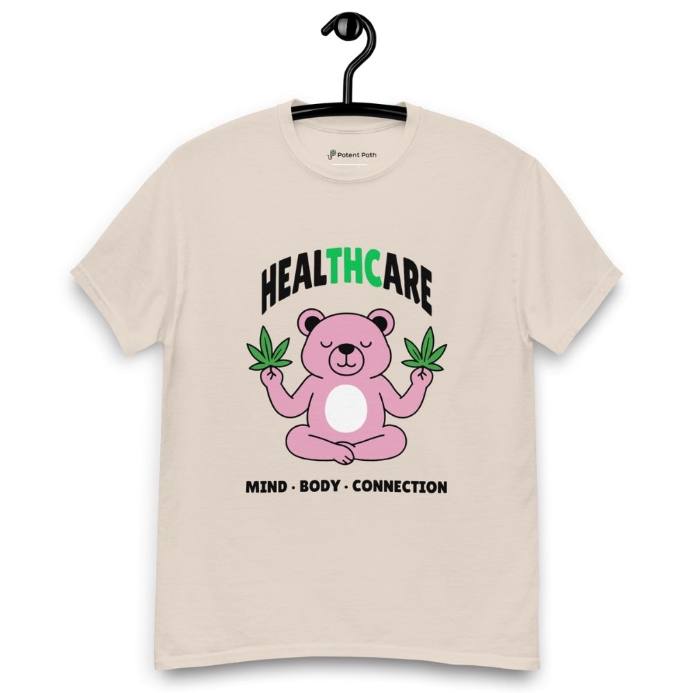 Beige T-shirt on a hanger featuring a pink cartoon bear sitting in a meditative pose and holding two cannabis leaves, with the word "HEALTHCARE" above, with the words "THC" highlighted in green and the phrase "MIND, BODY, CONNECTION" below