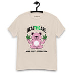 Beige T-shirt on a hanger featuring a pink cartoon bear sitting in a meditative pose and holding two cannabis leaves, with the word "HEALTHCARE" above, with the words "THC" highlighted in green and the phrase "MIND, BODY, CONNECTION" below