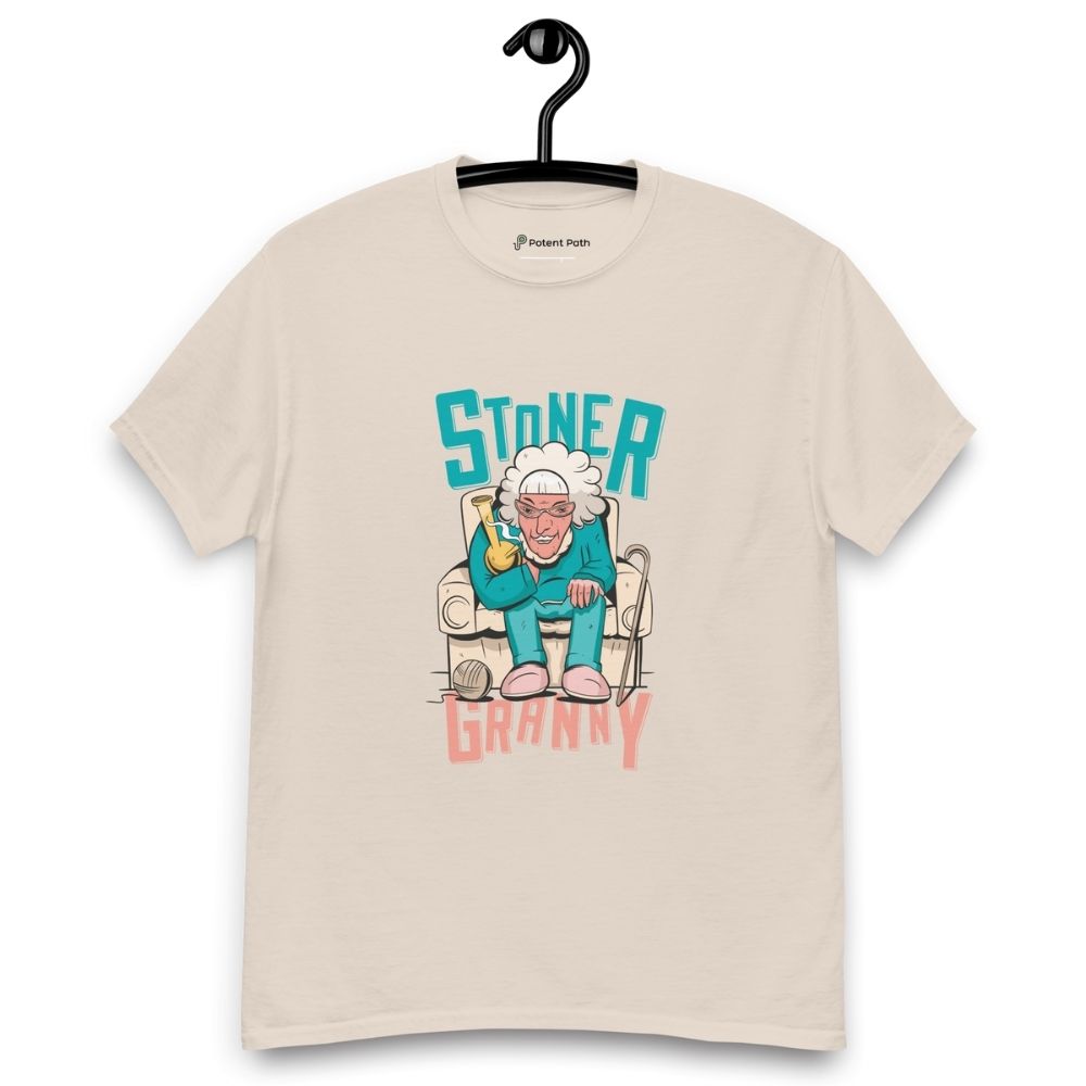 Beige T-shirt on a hanger featuring a cartoon illustration of an elderly woman in a teal tracksuit sitting in an armchair, holding a yellow bong, with the text "STONER GRANNY" above and below her.