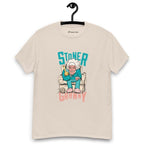 Beige T-shirt on a hanger featuring a cartoon illustration of an elderly woman in a teal tracksuit sitting in an armchair, holding a yellow bong, with the text "STONER GRANNY" above and below her.