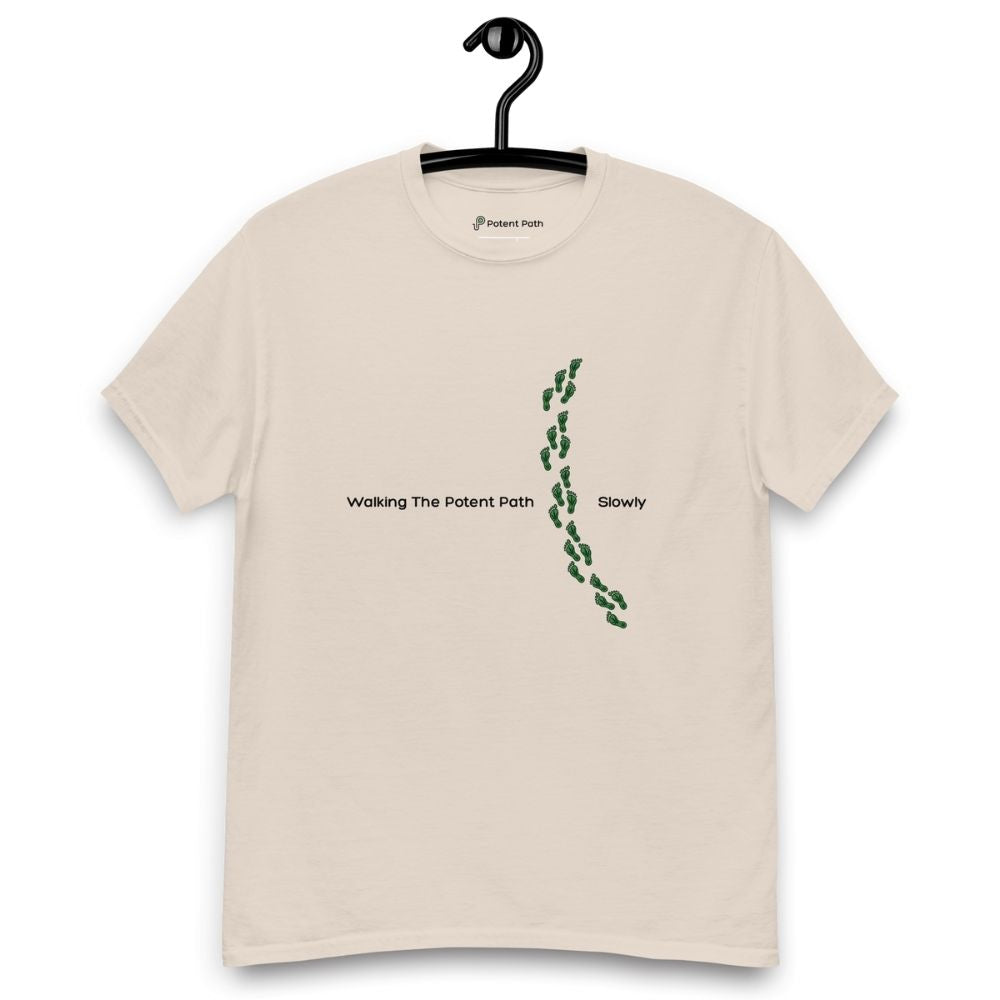 Beige t-shirt on a hanger featuring the phrase "Walking The Potent Path Slowly" in white text across the chest, with a vertical trail of small green footprints with cannabis leaves inside between "Path" and "Slowly"