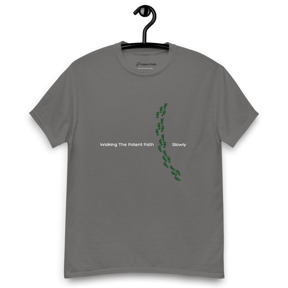Charcoal grey t-shirt on a hanger featuring the phrase "Walking The Potent Path Slowly" in white text across the chest, with a vertical trail of small green footprints with cannabis leaves inside between "Path" and "Slowly"