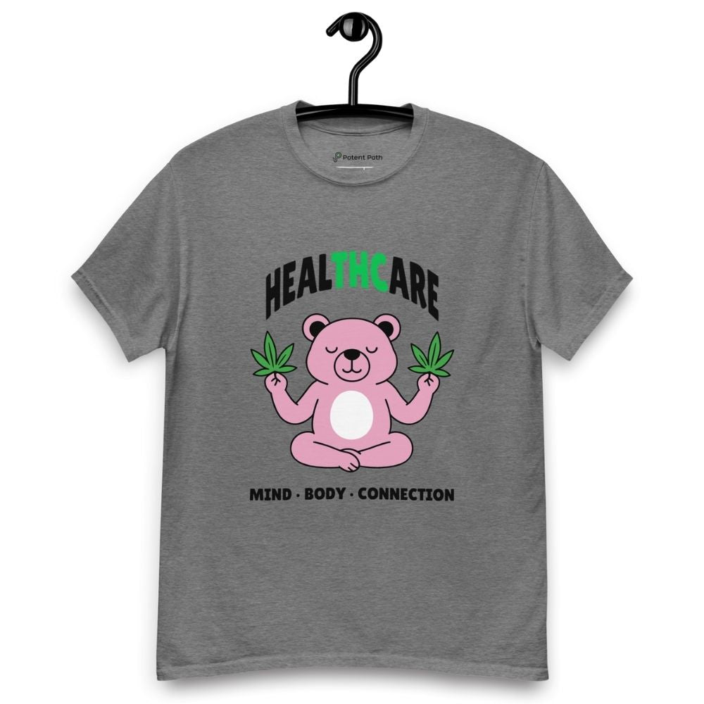 Grey T-shirt on a hanger featuring a pink cartoon bear sitting in a meditative pose and holding two cannabis leaves, with the word "HEALTHCARE" above, with the words "THC" highlighted in green and the phrase "MIND, BODY, CONNECTION" below