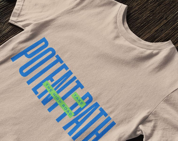 Close up of a beige t-shirt on a wooden surface with a print on the shirt which has large blue text that reads "POTENT PATH," with a small green overlay in the centre containing the words "TAKE THE HIGH ROAD"