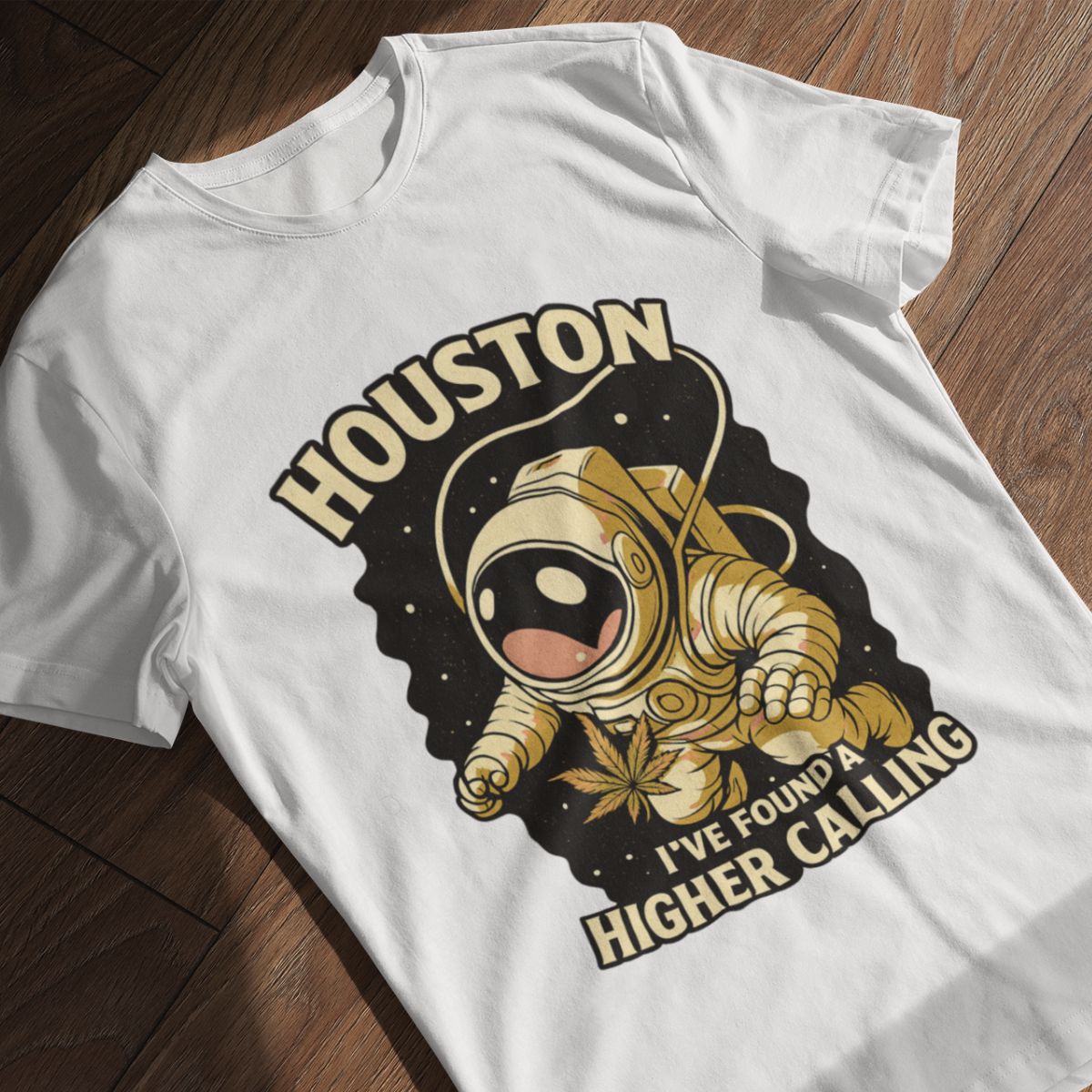 Light grey T-shirt laid flat on a wooden surface, featuring a cartoon astronaut floating in space while holding a cannabis leaf. The design includes the text "HOUSTON" at the top and "I'VE FOUND A HIGHER CALLING" at the bottom