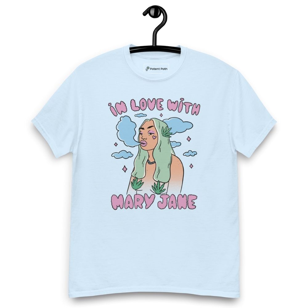 Light blue T-shirt on a hanger featuring a cartoon woman with long green hair, cannabis leaves covering her breasts and smoke clouds behind her, with the text "IN LOVE WITH MARY JANE" above and "MARY JANE" below the illustration
