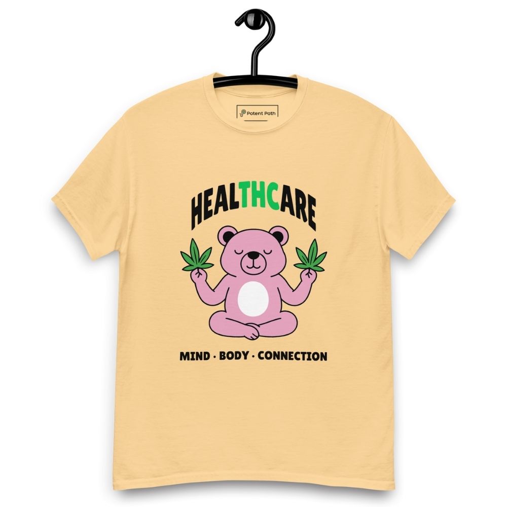 Light yellow T-shirt on a hanger featuring a pink cartoon bear sitting in a meditative pose and holding two cannabis leaves, with the word "HEALTHCARE" above, with the words "THC" highlighted in green and the phrase "MIND, BODY, CONNECTION" below