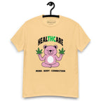 Light yellow T-shirt on a hanger featuring a pink cartoon bear sitting in a meditative pose and holding two cannabis leaves, with the word "HEALTHCARE" above, with the words "THC" highlighted in green and the phrase "MIND, BODY, CONNECTION" below