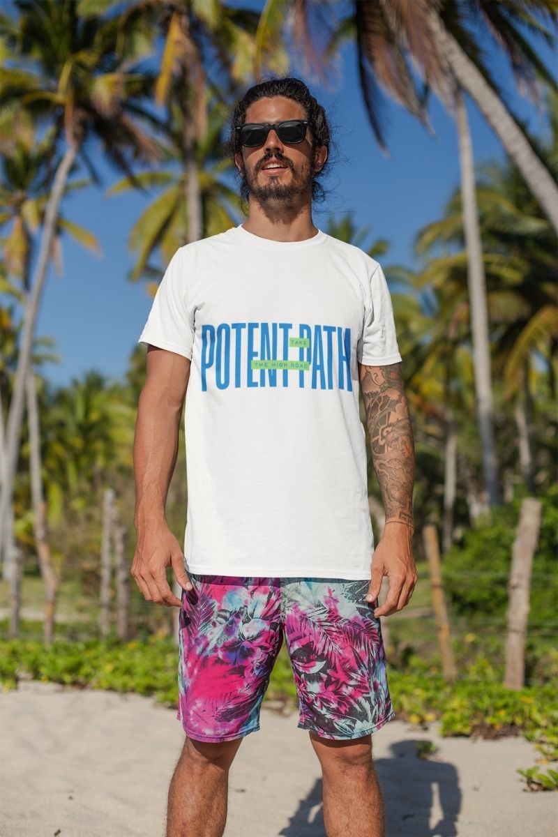 A man wearing sunglasses and a white Potent Path T-shirt, standing on a beach with palm trees behind him, dressed in tie-dye shorts under a bright blue sky