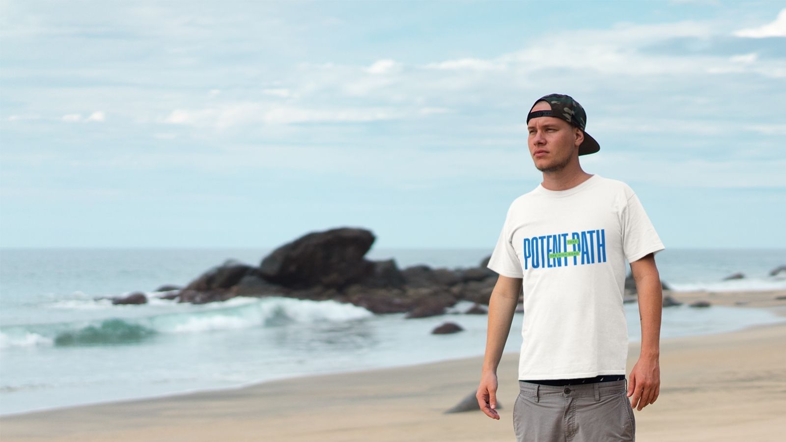 Man standing on a sandy beach wearing a white Potent Path T-shirt, grey shorts, and a backward cap, with ocean waves and rocky shoreline in the background under a cloudy sky