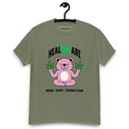 Military green T-shirt on a hanger featuring a pink cartoon bear sitting in a meditative pose and holding two cannabis leaves, with the word "HEALTHCARE" above, with the words "THC" highlighted in green and the phrase "MIND, BODY, CONNECTION" below