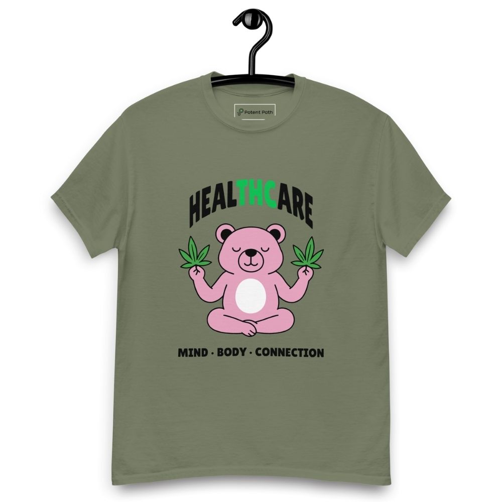 Military green T-shirt on a hanger featuring a pink cartoon bear sitting in a meditative pose and holding two cannabis leaves, with the word "HEALTHCARE" above, with the words "THC" highlighted in green and the phrase "MIND, BODY, CONNECTION" below