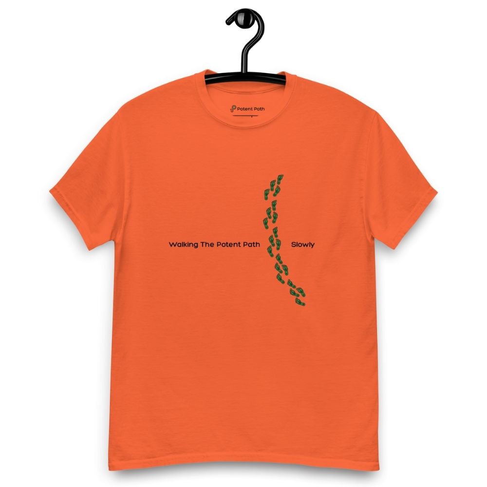 Orange t-shirt on a hanger featuring the phrase "Walking The Potent Path Slowly" in white text across the chest, with a vertical trail of small green footprints with cannabis leaves inside between "Path" and "Slowly"
