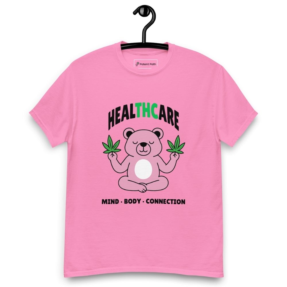 Pink T-shirt on a hanger featuring a pink cartoon bear sitting in a meditative pose and holding two cannabis leaves, with the word "HEALTHCARE" above, with the words "THC" highlighted in green and the phrase "MIND, BODY, CONNECTION" below