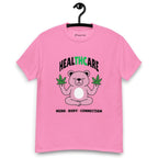 Pink T-shirt on a hanger featuring a pink cartoon bear sitting in a meditative pose and holding two cannabis leaves, with the word "HEALTHCARE" above, with the words "THC" highlighted in green and the phrase "MIND, BODY, CONNECTION" below