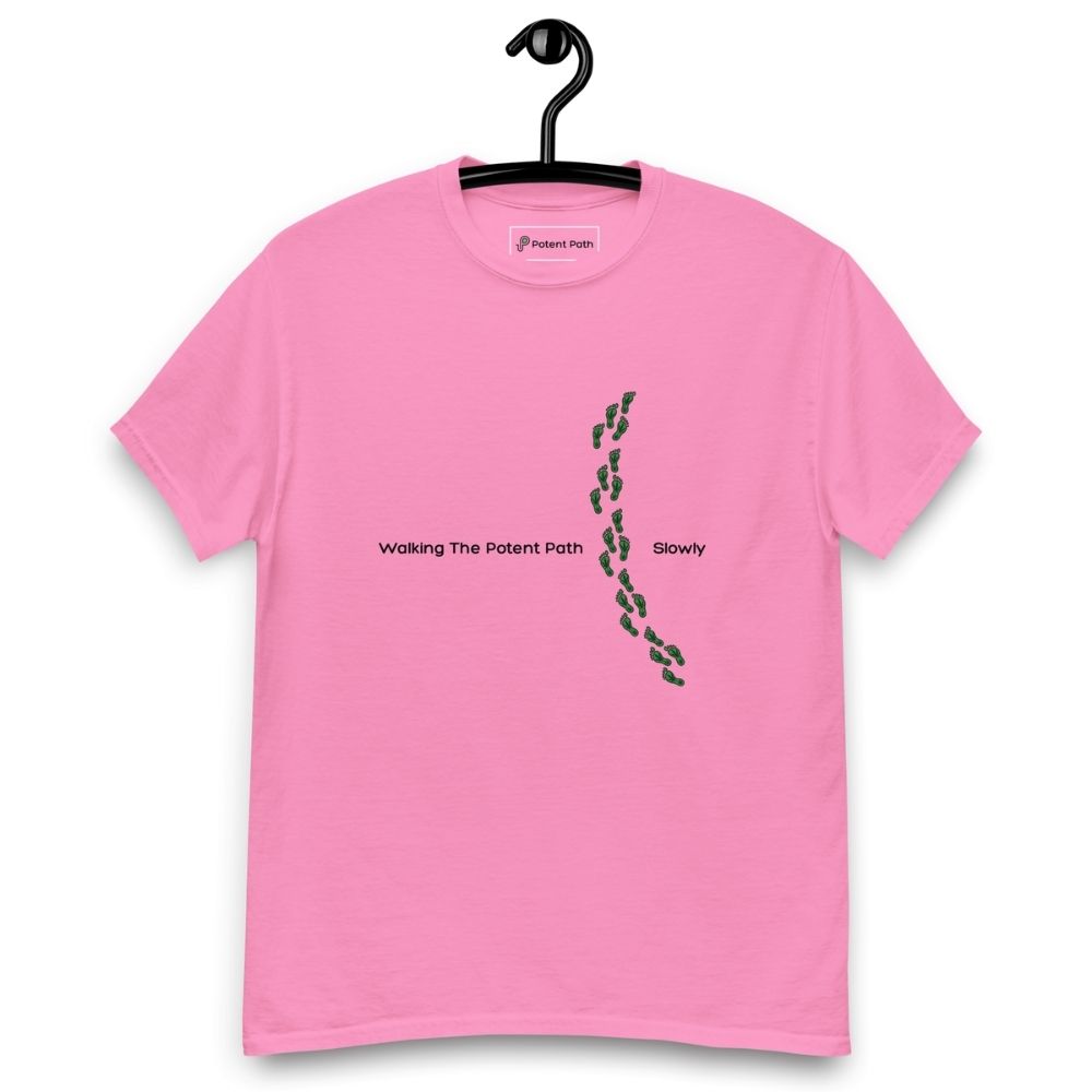 Pink t-shirt on a hanger featuring the phrase "Walking The Potent Path Slowly" in white text across the chest, with a vertical trail of small green footprints with cannabis leaves inside between "Path" and "Slowly"