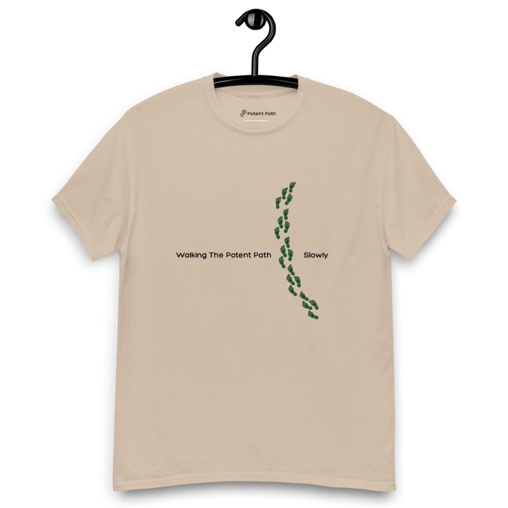 Sandy colour t-shirt on a hanger featuring the phrase "Walking The Potent Path Slowly" in white text across the chest, with a vertical trail of small green footprints with cannabis leaves inside between "Path" and "Slowly"
