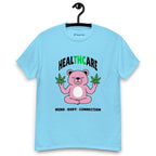 Sky blue T-shirt on a hanger featuring a pink cartoon bear sitting in a meditative pose and holding two cannabis leaves, with the word "HEALTHCARE" above, with the words "THC" highlighted in green and the phrase "MIND, BODY, CONNECTION" below