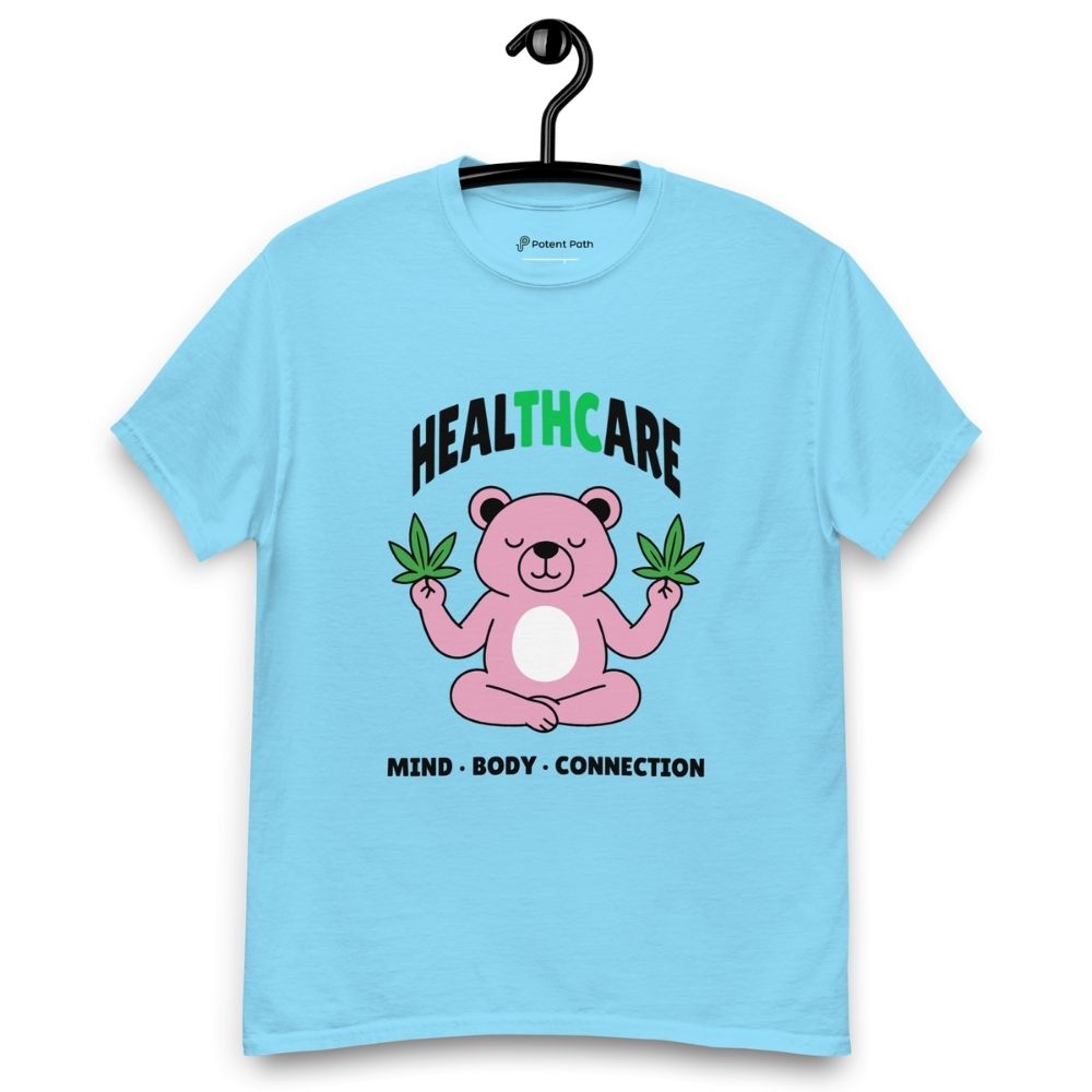 Sky blue T-shirt on a hanger featuring a pink cartoon bear sitting in a meditative pose and holding two cannabis leaves, with the word "HEALTHCARE" above, with the words "THC" highlighted in green and the phrase "MIND, BODY, CONNECTION" below