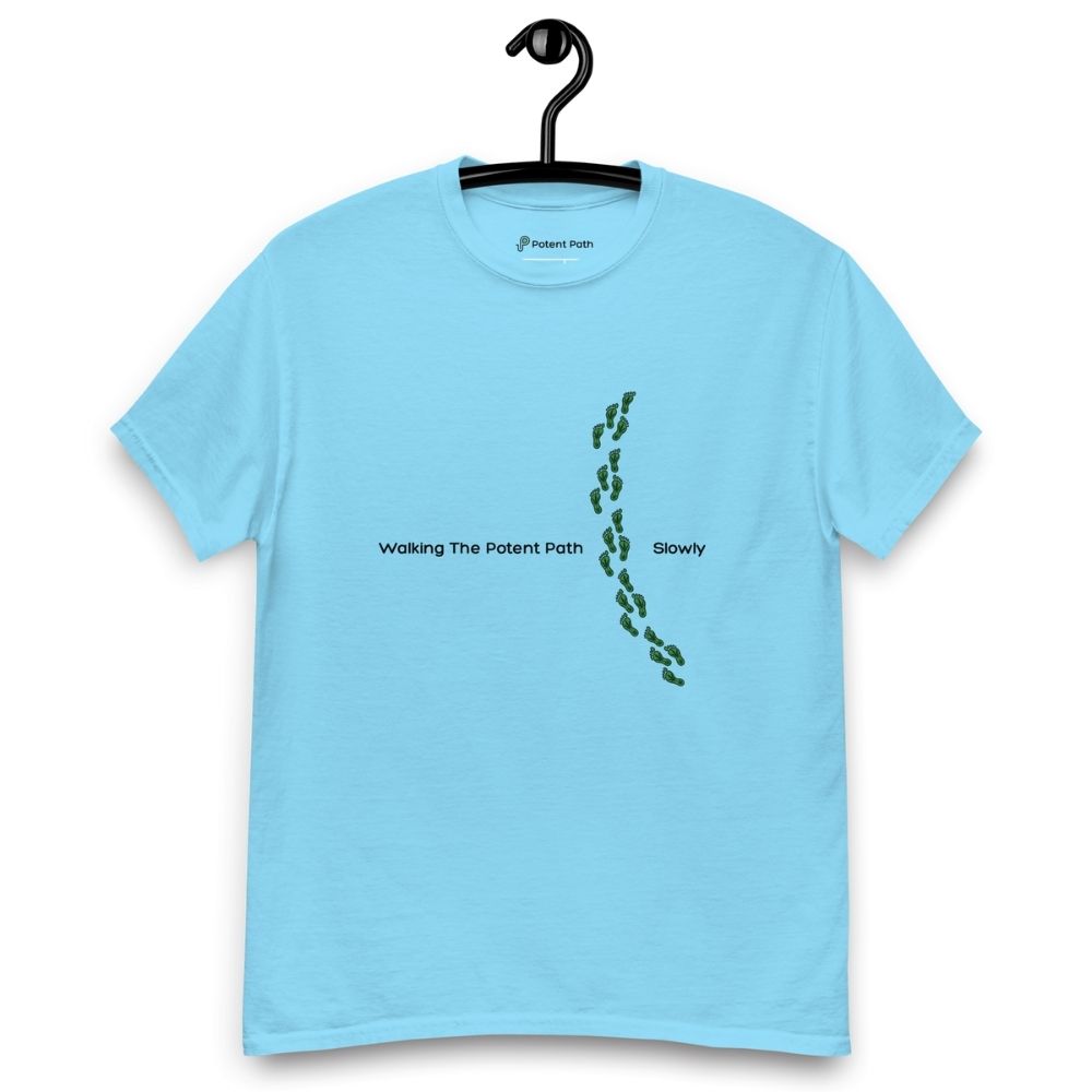 Sky blue t-shirt on a hanger featuring the phrase "Walking The Potent Path Slowly" in white text across the chest, with a vertical trail of small green footprints with cannabis leaves inside between "Path" and "Slowly"