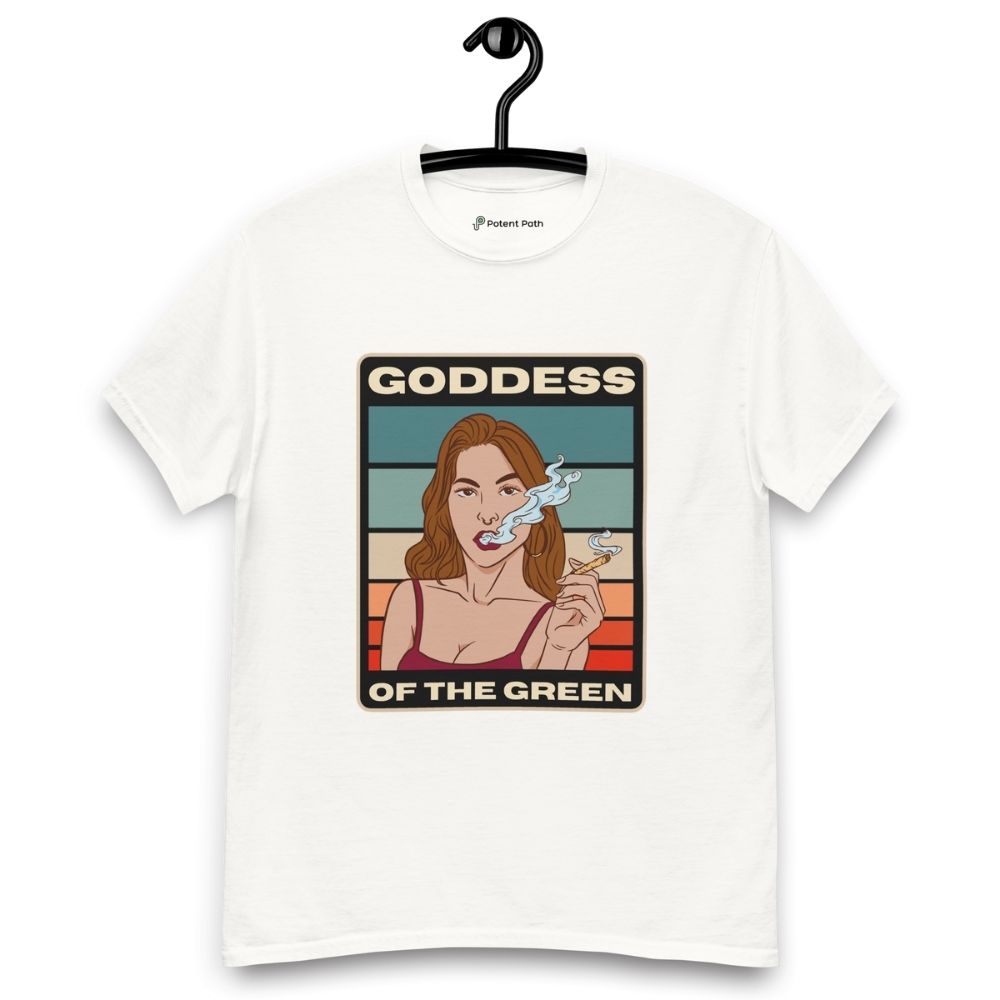 White T-shirt on a hanger featuring a retro-style illustration of a woman smoking a joint, set against colourful horizontal stripes, with the text "GODDESS" above and "OF THE GREEN" below the artwork