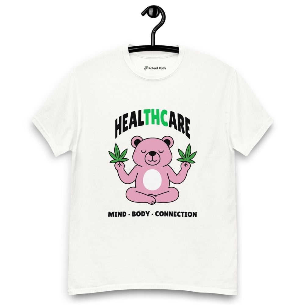 White T-shirt on a hanger featuring a pink cartoon bear sitting in a meditative pose and holding two cannabis leaves, with the word "HEALTHCARE" above, with the words "THC" highlighted in green and the phrase "MIND, BODY, CONNECTION" below