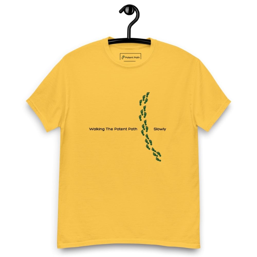 Yellow t-shirt on a hanger featuring the phrase "Walking The Potent Path Slowly" in white text across the chest, with a vertical trail of small green footprints with cannabis leaves inside between "Path" and "Slowly"